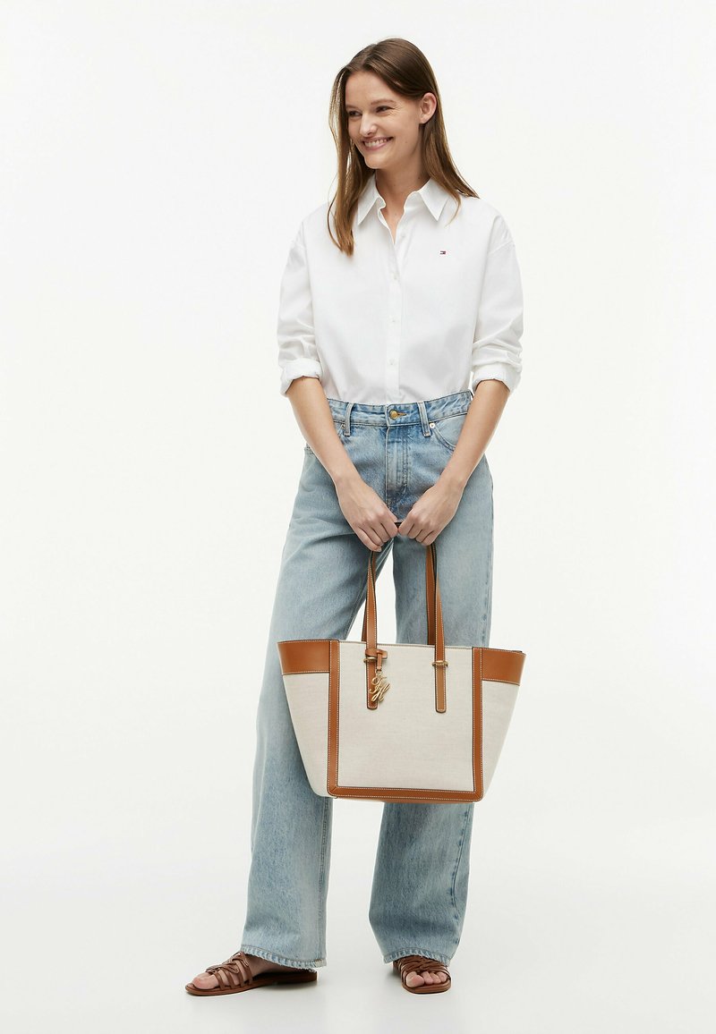 Femme en chemise blanche boutonnée et jean bleu clair tenant un sac cabas beige avec garniture et anses en cuir marron, souriante et debout.