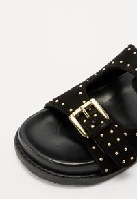 Sandalo nero in pelle scamosciata con borchie dorate, fascia larga e grande fibbia metallica. Presenta una soletta nera testurizzata per garantire aderenza.