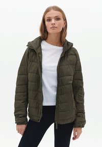 Olijfgroene pufferjack met een geborduurd ontwerp, ritsluiting en verstelbare capuchon. Draag het over een wit shirt en combineer het met zwarte leggings.