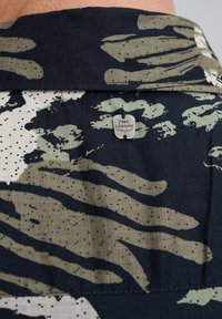 Camisa de manga corta con un patrón de camuflaje en tonos verde oscuro, beige y negro, con una etiqueta plateada "PME Legend" en el cuello.