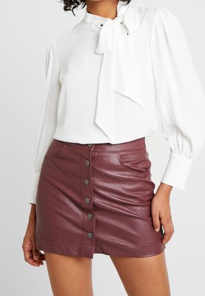 Mujer que lleva una blusa blanca de manga larga con lazo en el cuello y una minifalda de cuero burdeos con botones delante.