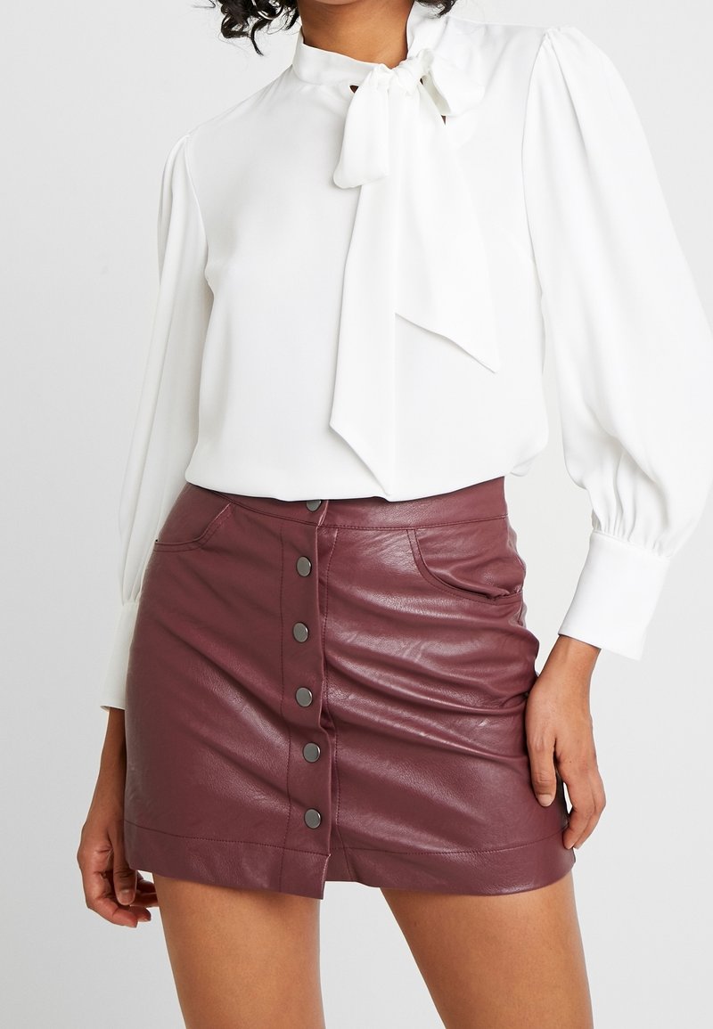 Femme portant un chemisier blanc à manches longues avec un nœud au col et une mini-jupe en cuir bordeaux boutonnée devant.