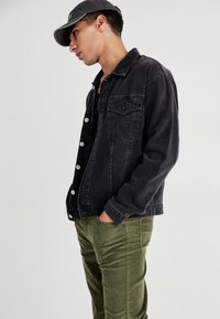Black denim trucker jacket with a front pocket, silver buttons, worn over a black shirt, paired with olive green corduroy pants.