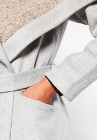 Cappotto in misto lana grigio con colletto beige testurizzato, dotato di vita con cintura, ampie rever e tasca laterale con etichetta.