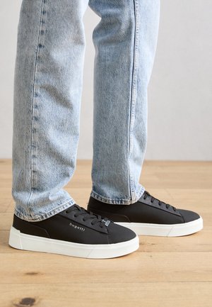 Zapatillas negras con un acabado liso, suela de goma blanca y un sutil logotipo en el lateral, combinadas con jeans de mezclilla azul claro.