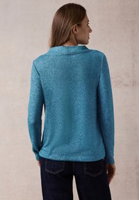 Helle türkisfarbene langärmlige Bluse aus strukturiertem Stoff mit einem spitzen Kragen, kombiniert mit dunkelblauen Jeans.