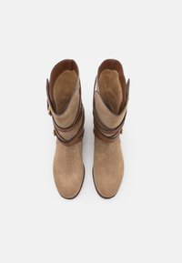Bottes en daim tan avec des sangles en cuir marron, un bout arrondi et une surface texturée. Des accents en matériel doré sont visibles.