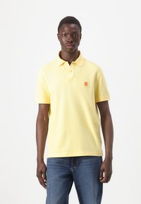 Žlté polo tričko z mäkkého materiálu, s klasickým golierom a trojradovým gombíkom, s malým oranžovým logom na hrudi.