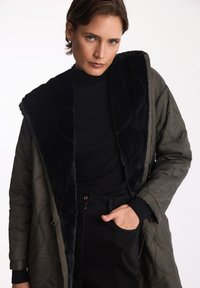 Kurtka w oliwkowym kolorze, pikowana, z czarnym kołnierzem z sztucznego futra, z wzorem w diamenty, bocznymi kieszeniami oraz luźnym krojem, noszona na czarnej bluzce i spodniach.