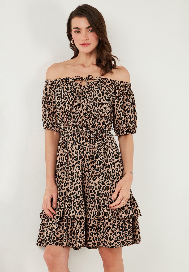Off-the-shoulder leopard print dress in beige with black spots. Soft fabric, elastic cuffs, fitted waist, and tiered frill hem.