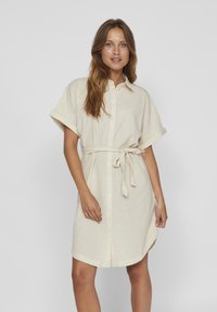 Svijetlosmeđa lanena haljina sa kratkim rukavima, dugmićima sprijeda i vezivanjem u struku. Ima ovratnik i opušteni kroj. Mekana tekstura.