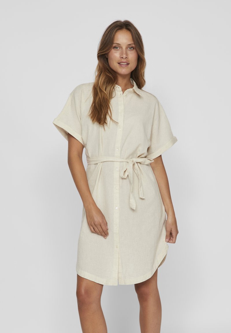 Svijetlosmeđa lanena haljina sa kratkim rukavima, dugmićima sprijeda i vezivanjem u struku. Ima ovratnik i opušteni kroj. Mekana tekstura.