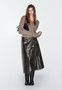 Frau trägt einen braunen Strickpullover, einen dunkelbraunen Leder-Midirock, schwarze kniehohe Stiefel und hält eine kleine schwarze Handtasche.