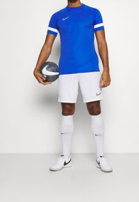 Blue athletic shirt with white stripes, white shorts, and knee-high white socks. Holding a black and gray soccer ball, wearing white sneakers.