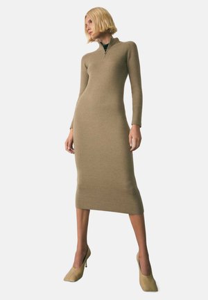 Robe longue et ajustée en tissu côtelé, de couleur marron clair, dotée d'un col zippé, de manches longues et d'une longueur midi, associée à des talons beiges.