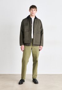 Quilted olivgrüne Jacke mit schwarzem Kragen, auffälligen Knöpfen und Brusttaschen, kombiniert mit olivfarbenen Hosen und schwarzen Schuhen.