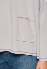 Hellgraues Oberteil aus strukturiertem Stoff, mit einer Brusttasche und silberner Dekostich entlang des Saums und der Ärmelsäume.