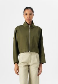 Chaqueta verde oliva recortada con un cuello alto, cierre frontal y dos grandes bolsillos; fabricada en un tejido suave y ligero.