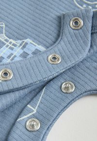 Primer plano de tela azul acanalada con botones a presión plateados parcialmente abrochados en una prenda con un diseño impreso claro.