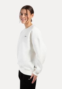 Jersey de cuello redondo blanco hecho de tela suave con puños y dobladillo acanalados, presenta un pequeño logo en el pecho y un corte relajado.