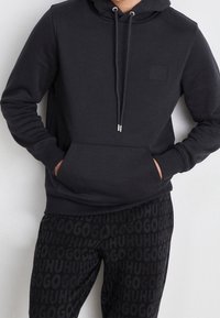 Hoodie noir avec une poche kangourou, capuche avec cordon de serrage, et un petit écusson. Associé à un pantalon de survêtement noir avec un logo en texte répété.