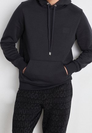 Sudadera negra con bolsillo tipo canguro, capucha con cordón y un pequeño parche. Combinada con pantalones de chándal negros que presentan un logo de texto repetido.