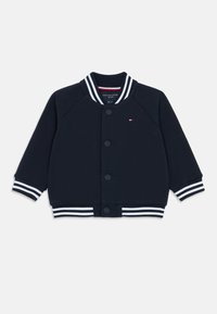 Námořnická modrá varsity bunda se bílými pruhovanými ribovými límcem a manžetami, zapínání na knoflíky a nenápadným logem na levé hrudi.