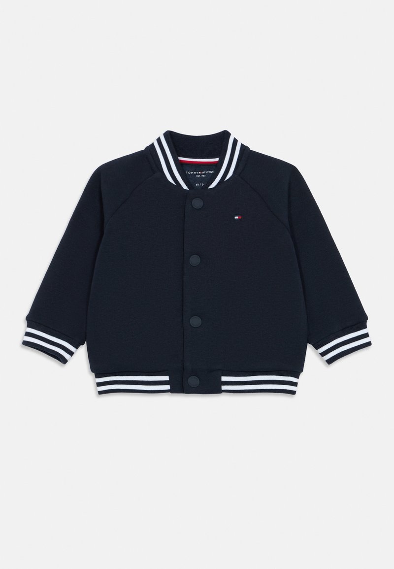 Námořnická modrá varsity bunda se bílými pruhovanými ribovými límcem a manžetami, zapínání na knoflíky a nenápadným logem na levé hrudi.