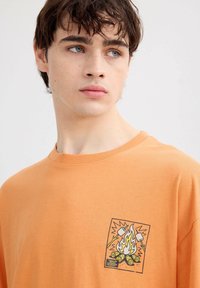 Joven de piel clara y cabello oscuro que lleva una camiseta naranja con un gráfico de una hoguera y malvaviscos asándose.