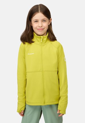 Ragazza giovane con capelli castani lisci, indossa una giacca lime verde Mammut con cerniera e pantaloni verde chiaro, sorride guardando verso avanti.