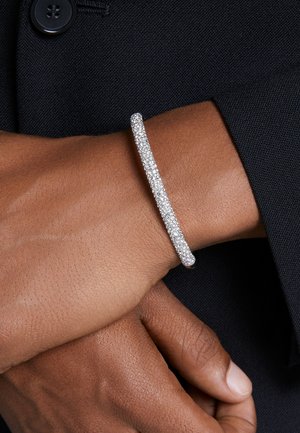 Silberarmband mit geschwungenem Design, besetzt mit mehreren kleinen funkelnden Steinen, getragen an einem Handgelenk vor einem dunklen Stoffhintergrund.