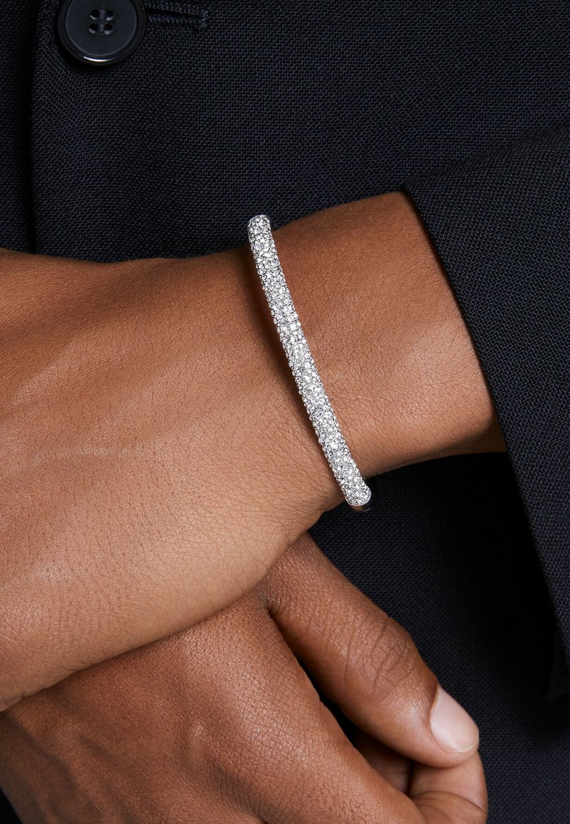 Pulsera de plata con un diseño curvado, adornada con múltiples pequeñas piedras brillantes, llevada en una muñeca sobre un fondo de tela oscura.