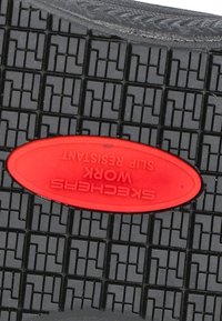Suela de zapato antideslizante negra con un patrón texturizado; presenta una etiqueta ovalada roja con "SKECHERS WORK" en relieve.