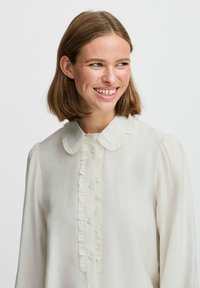 Weiße Bluse mit einer Rüschencollage und einer Knopfleiste auf der Vorderseite. Lange Ärmel mit leichtem Puff an den Schultern. Glatte Stofftextur.