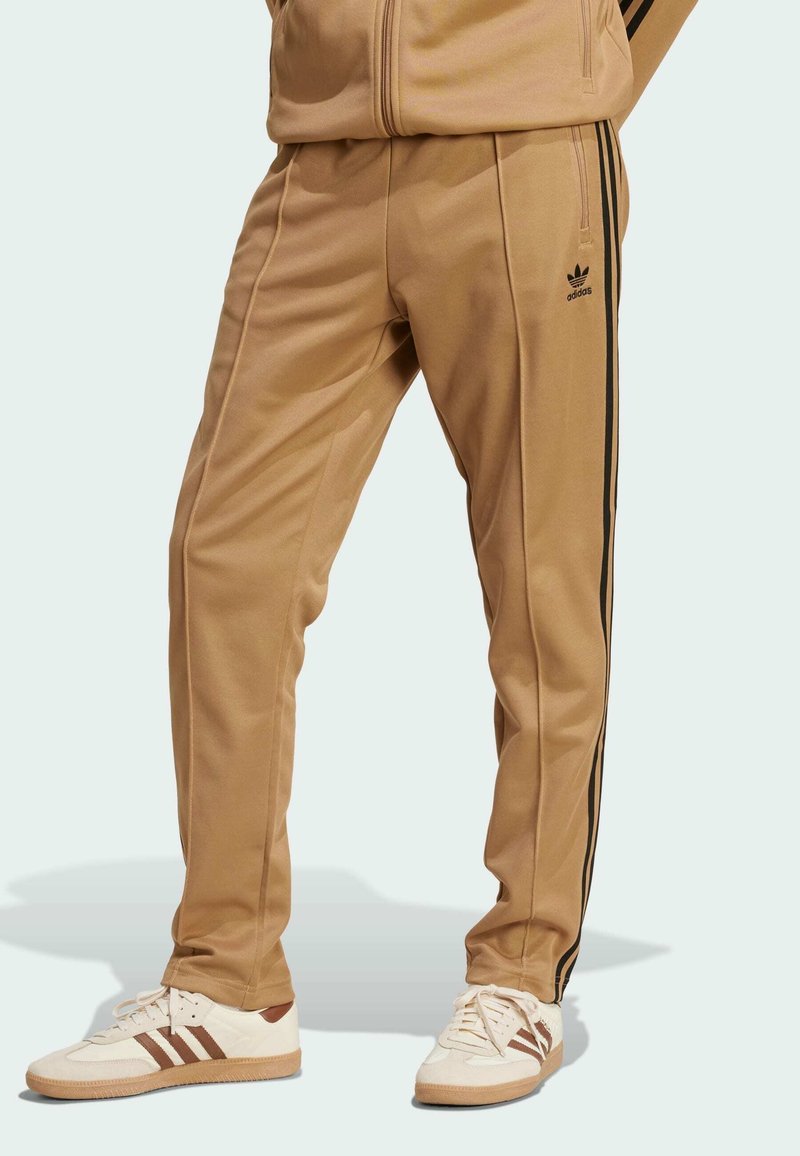 Brown athletic pants with black side stripes, made from smooth fabric. Paired with white shoes featuring brown accents and stripes.