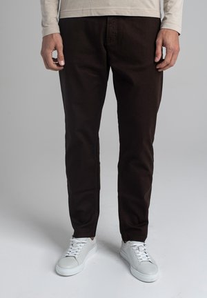 Mężczyzna ubrany w ciemnobrązowe spodnie, białe trampki i jasnobeżową koszulę z długim rękawem stoi na jednolitym szarym tle.