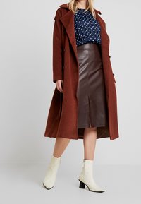 Lungo cappotto di lana color ruggine con bottoni neri, indossato sopra una blusa blu navy con pois bianchi e una gonna di pelle marrone. Stivaletti bianchi alla caviglia.