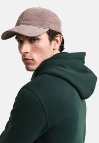 Gorra de pana marrón con visera curvada, combinada con una sudadera verde oscuro que presenta una gran capucha y una textura de tela suave.