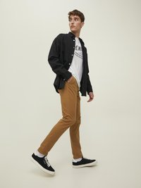 Czarna koszula zapinana na guziki na białym T-shircie z grafiką, beżowe spodnie slim-fit oraz czarne sneakersy z białą podeszwą, stojący na neutralnym tle.