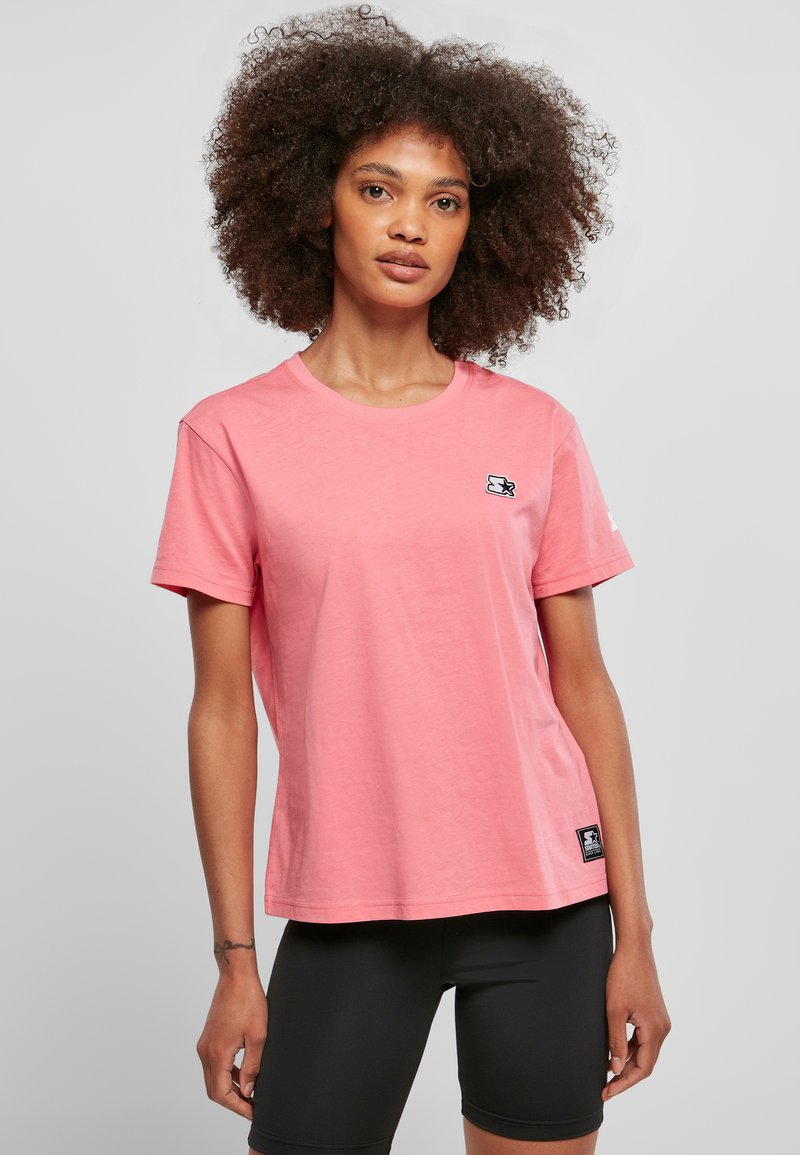 Tricou roz deschis din amestec de bumbac, cu mâneci scurte, decolteu rotund și o mică etichetă cu logo pe piept. Asortat cu pantaloni scurți negri.
