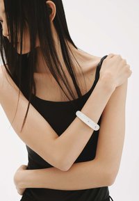 Pulsera blanca y brillante en un brazo desnudo. El brazo está posicionado sobre el pecho, y la persona lleva una camiseta sin mangas negra.