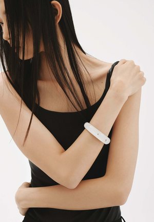 Pulsera blanca y brillante en un brazo desnudo. El brazo está posicionado sobre el pecho, y la persona lleva una camiseta sin mangas negra.