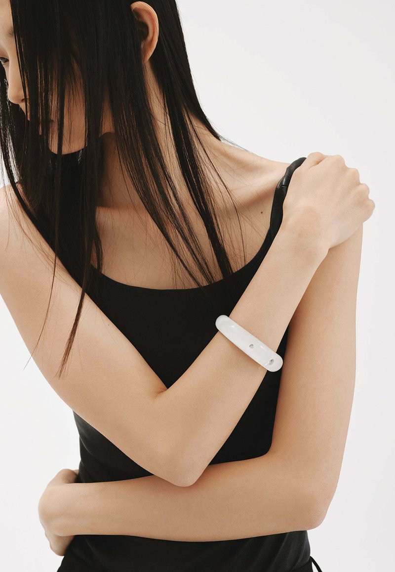 Pulsera blanca y brillante en un brazo desnudo. El brazo está posicionado sobre el pecho, y la persona lleva una camiseta sin mangas negra.