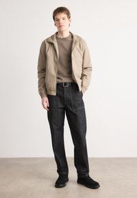 Giacca beige con polsini elasticizzati, maglione marrone e pantaloni di jeans scuri con tasche anteriori, abbinati a scarpe nere su uno sfondo neutro.