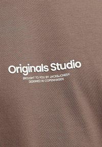 Brauner Stoff mit weißem Text, der "Originals Studio" sagt, gefolgt von kleinerem Text darunter, der die Marke und den Designursprung angibt.