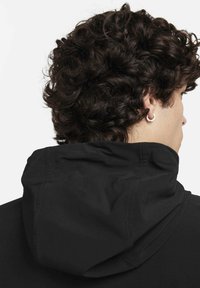 Zwarte capuchonjas met een gladde textuur, voorzien van een aangehechte capuchon en platte naden, gezien van achteren, met krullend haar en een zilveren oorbel.