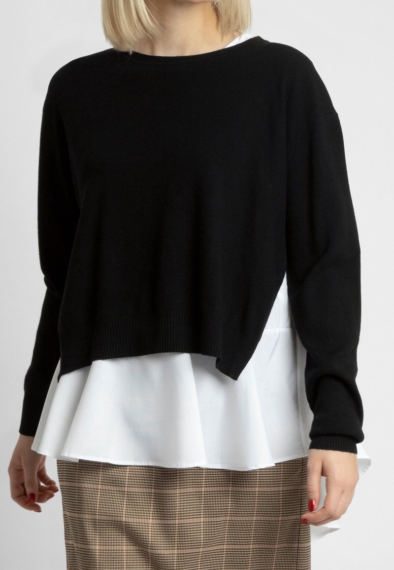 Femme porte un pull noir à manches longues superposé sur un chemisier blanc à ourlet évasé, associé à une jupe beige à carreaux, debout devant un fond uni.