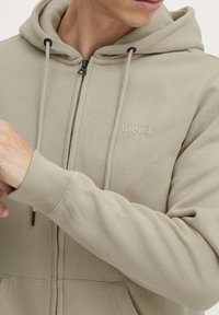 Felpa con zip beige caratterizzata da una texture morbida, cappuccio regolabile con lacci e tasche frontali, con un sottile logo "blend" sul petto.