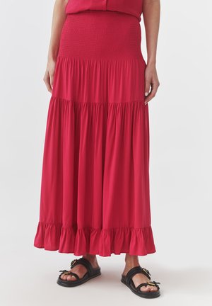 Maxi gonna rossa con vita arricciata, balze a strati e silhouette fluida. Indossata con sandali neri con accenti di hardware decorativo.