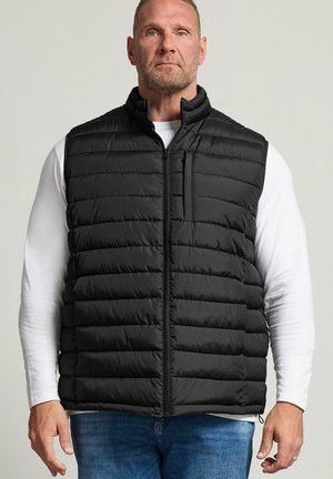 Homme portant une veste matelassée noire sur une chemise blanche à manches longues et un jean bleu sur fond uni.